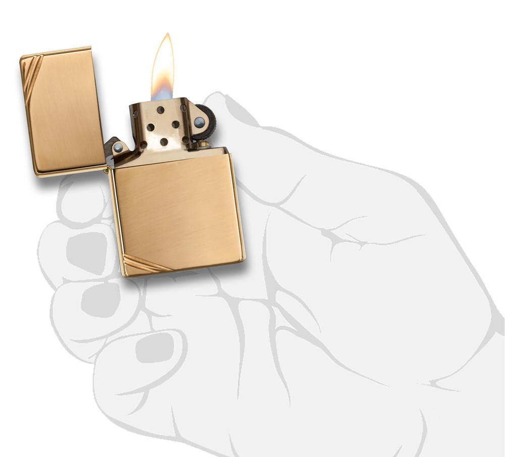 Vue de face briquet Zippo Vintage Brass High Polished avec des entailles décoratives aux deux coins, ouvert avec flamme dans une main stylisée